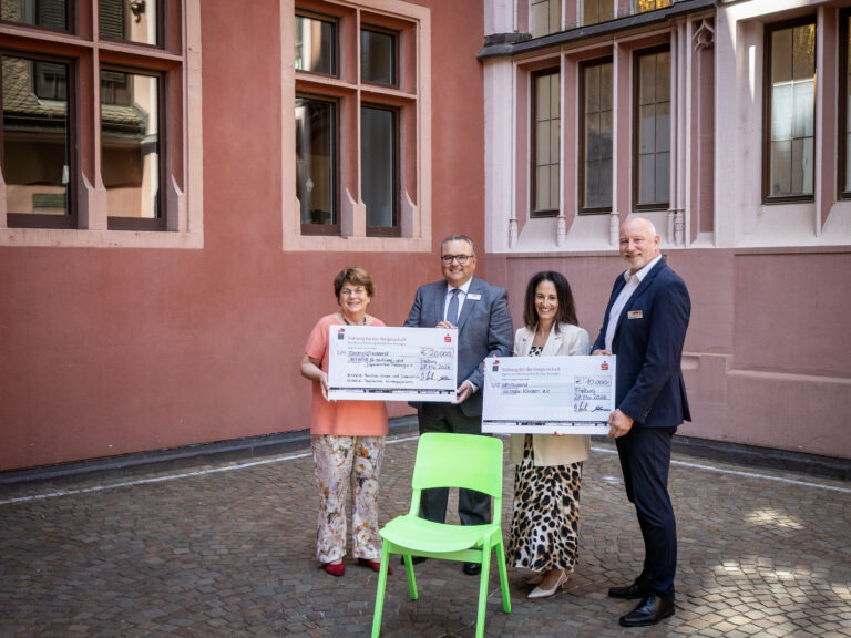 Group of four adults in smart clothing posing outdoors, holding oversized ceremonial checks in front of a pink building; a bright green chair is in the foreground.