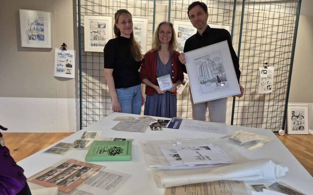 Three people pose behind a blue metal grid display with framed comic panels at an art exhibit; a table with papers and booklets is in the foreground.