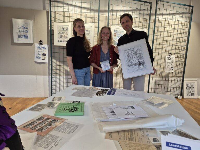 Three people pose behind a blue metal grid display with framed comic panels at an art exhibit; a table with papers and booklets is in the foreground.