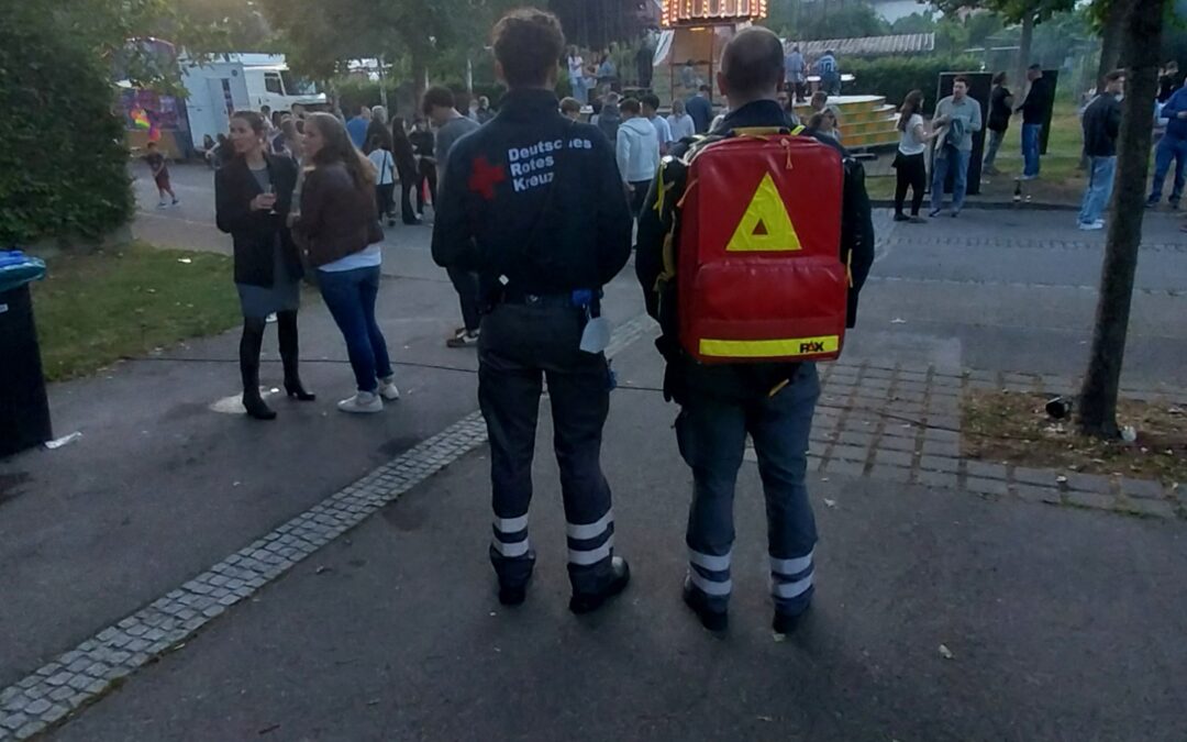 Two responders with reflective gear stand with their backs to the camera, a red backpack visible, as a lit carousel and church spire loom in the distance at a park festival.