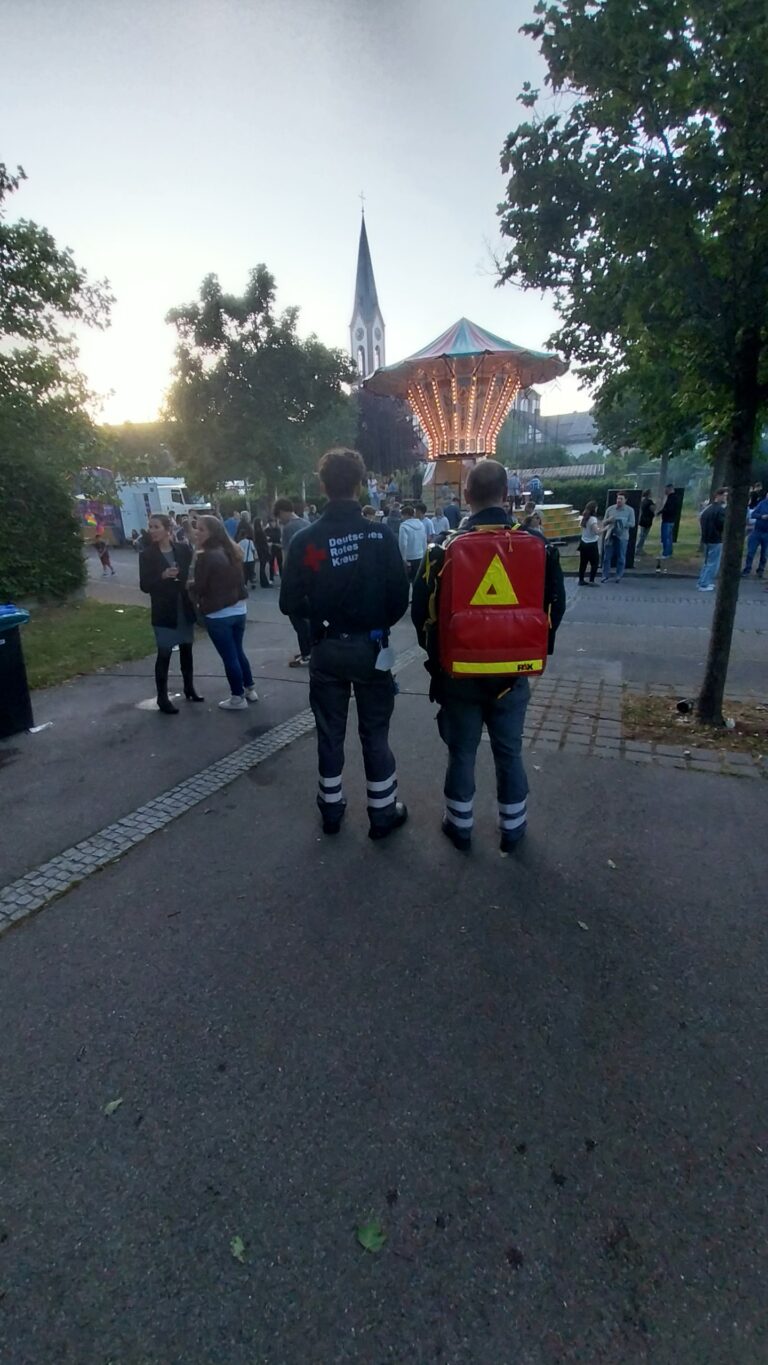Two responders with reflective gear stand with their backs to the camera, a red backpack visible, as a lit carousel and church spire loom in the distance at a park festival.