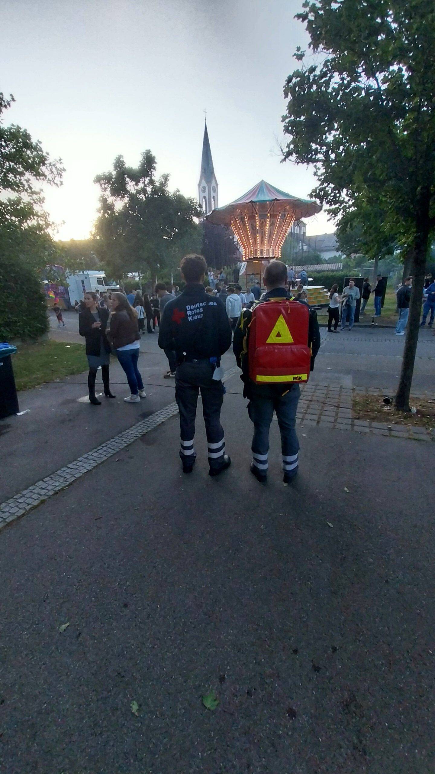 Two responders with reflective gear stand with their backs to the camera, a red backpack visible, as a lit carousel and church spire loom in the distance at a park festival.
