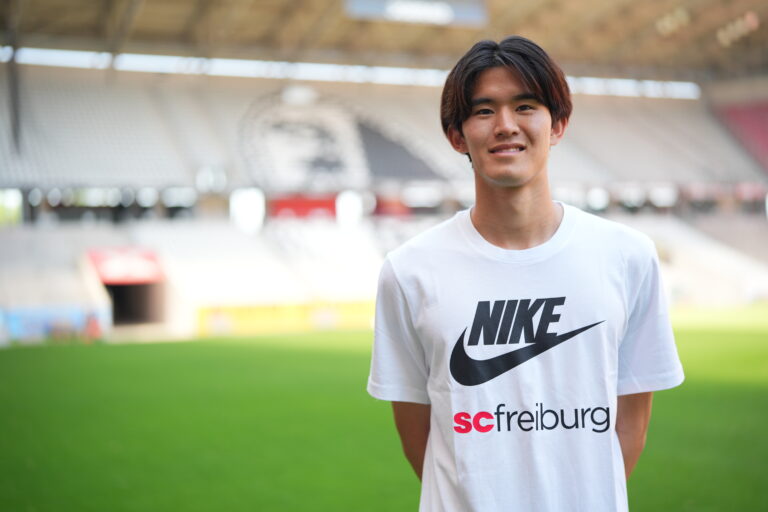 Smiling young man in a white Nike t-shirt stands on a soccer stadium field with blurred stands in the background.