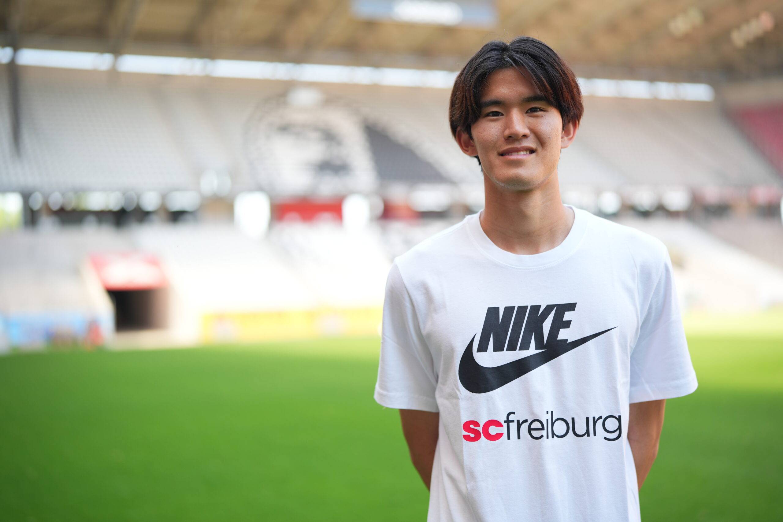 Smiling young man in a white Nike t-shirt stands on a soccer stadium field with blurred stands in the background.