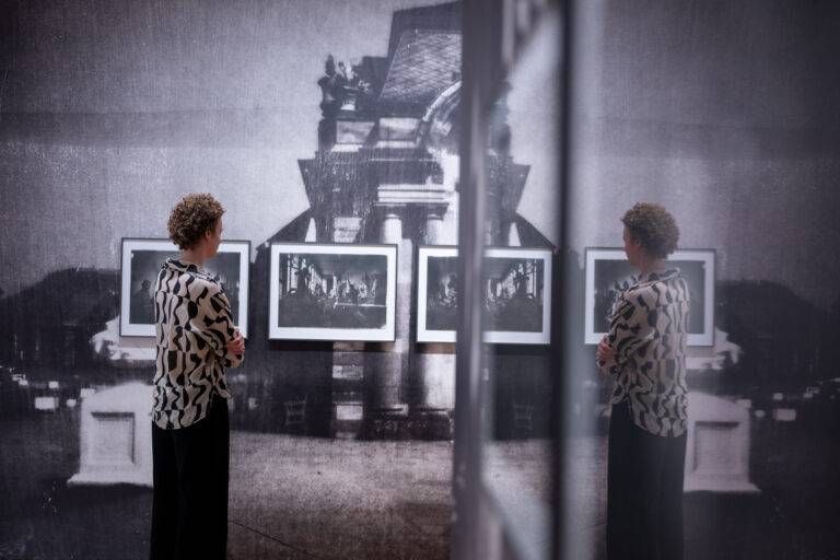 Person in a bold patterned shirt stands in a gallery, examining framed black-and-white photos on a wall; a reflective surface creates a duplicate view.