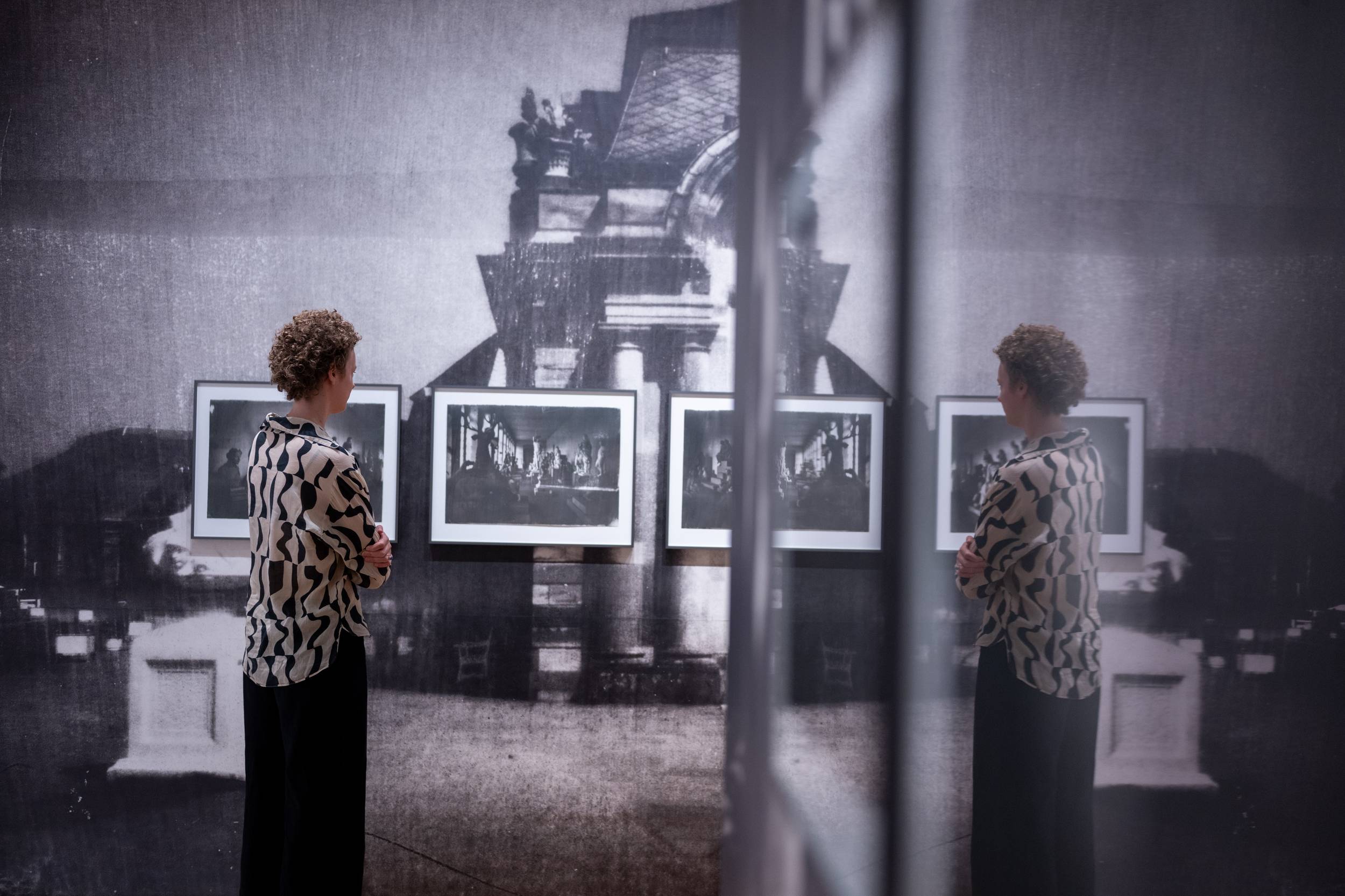 Person in a bold patterned shirt stands in a gallery, examining framed black-and-white photos on a wall; a reflective surface creates a duplicate view.