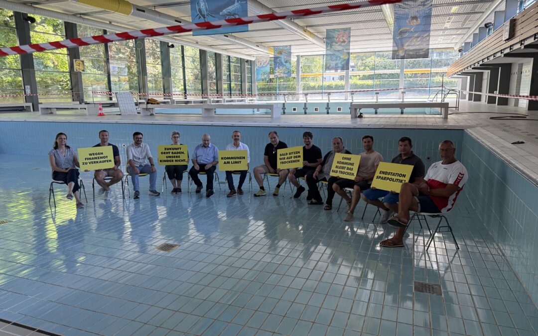 Group of people seated along the edge of an indoor swimming pool, each holding yellow protest signs with German slogans like 'Endstation Sparpolitik' or similar. Red tape marks off the area and banners hang from the ceiling.