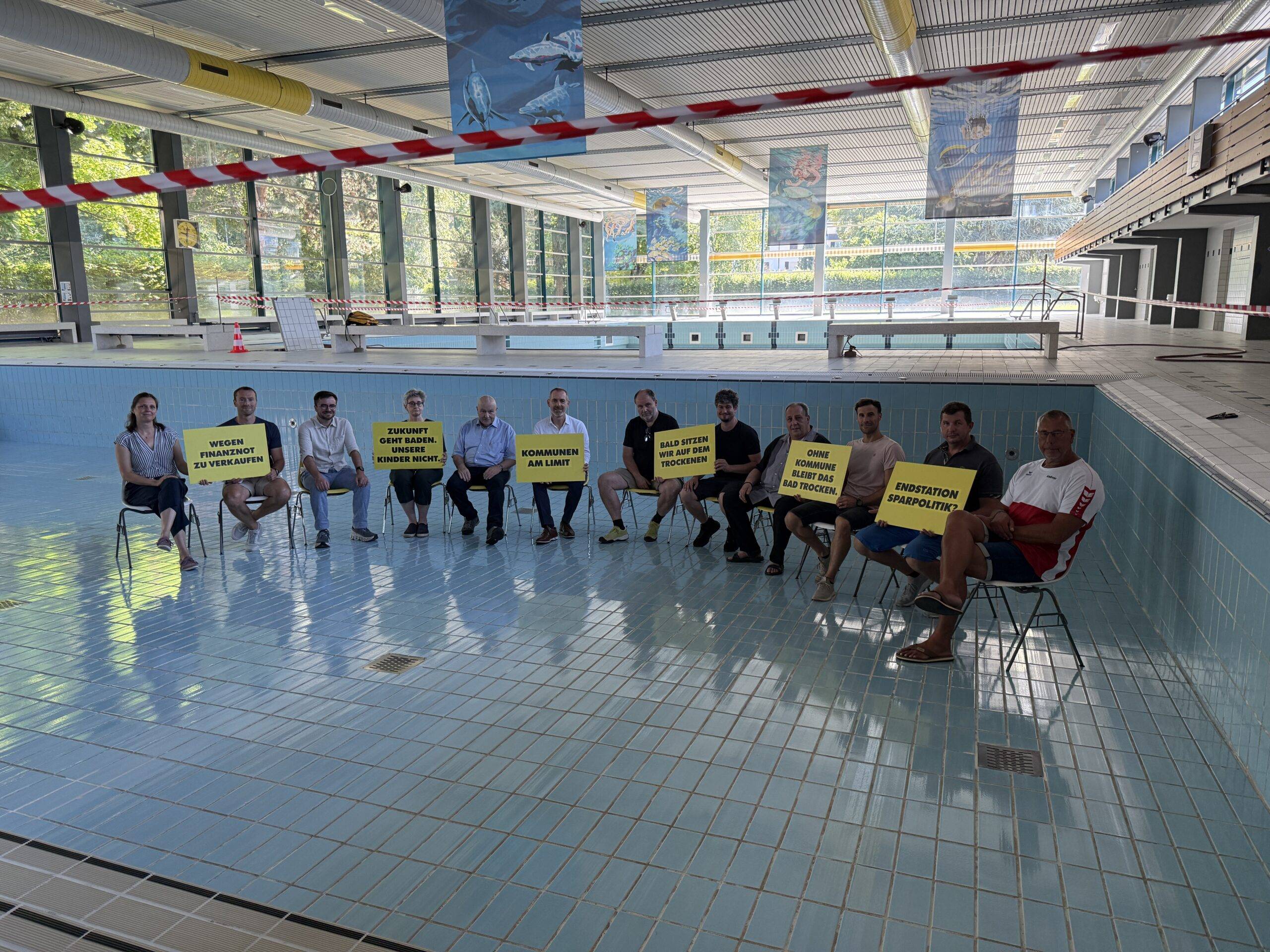Group of people seated along the edge of an indoor swimming pool, each holding yellow protest signs with German slogans like 'Endstation Sparpolitik' or similar. Red tape marks off the area and banners hang from the ceiling.
