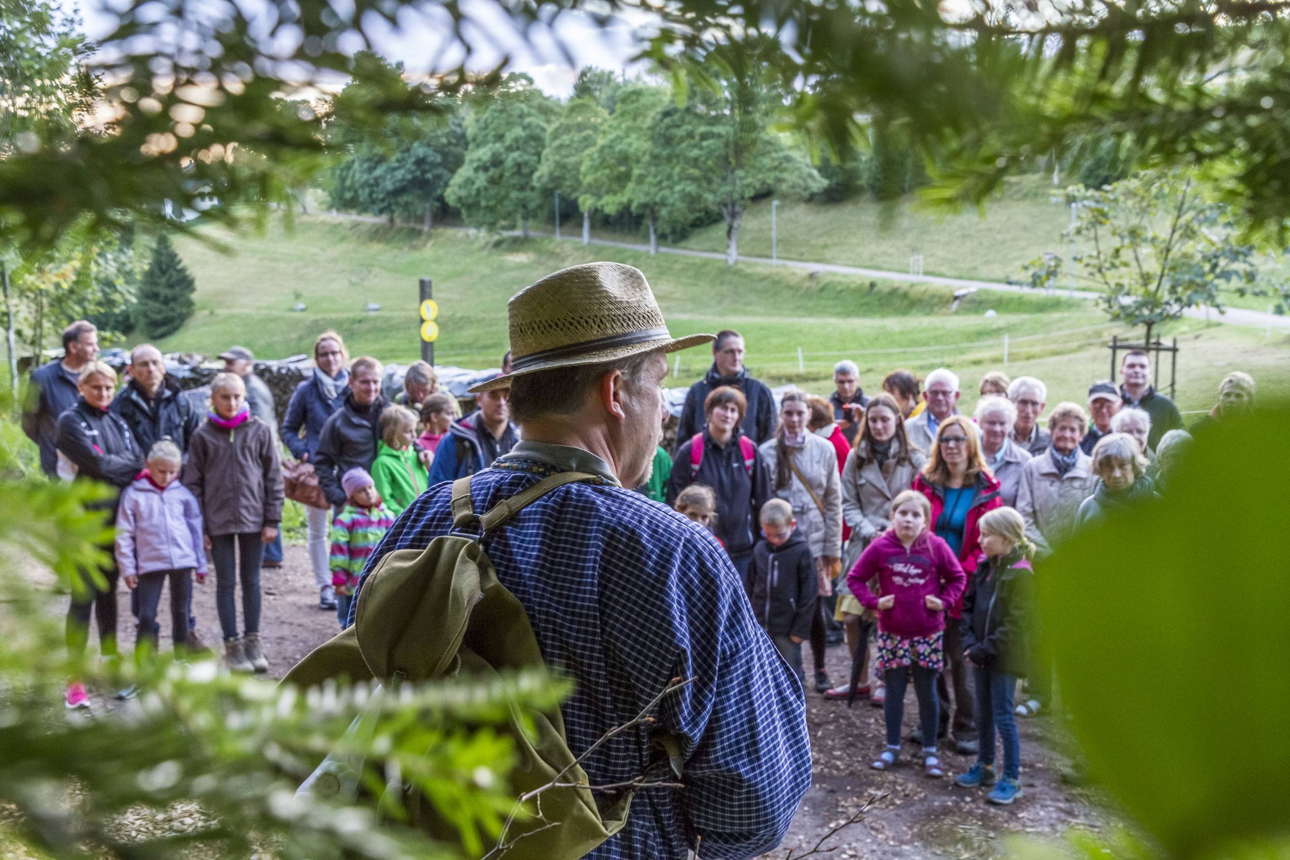 Man with a straw hat and backpack addressing a crowd in a park, with trees and a path in the background.