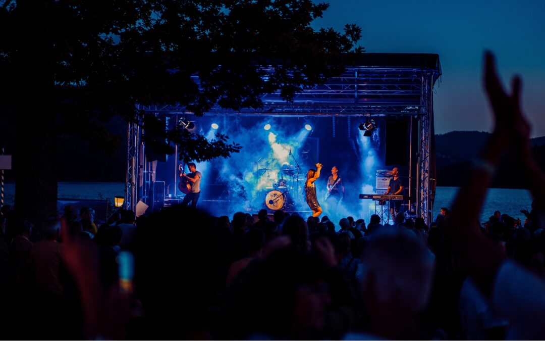 Nighttime outdoor concert on a lit stage with blue spotlights; the crowd raises hands in front of a lake.