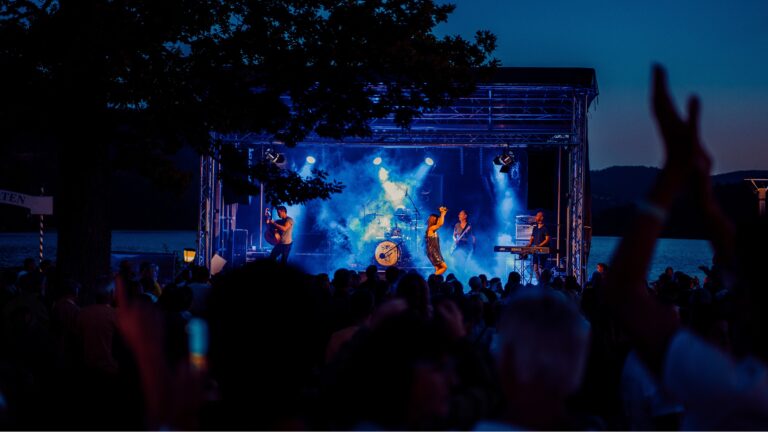 Nighttime outdoor concert on a lit stage with blue spotlights; the crowd raises hands in front of a lake.