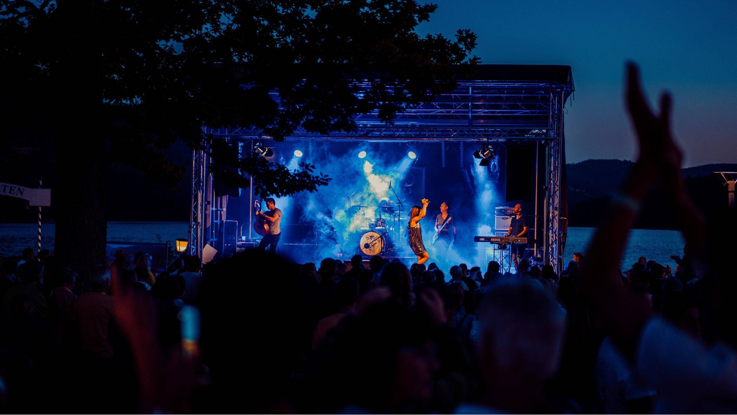 Nighttime outdoor concert on a lit stage with blue spotlights; the crowd raises hands in front of a lake.