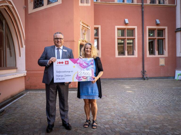 Two smiling adults in business attire hold an oversized cheque outdoors in front of a peach-colored building with arched windows and cobblestone ground.”](