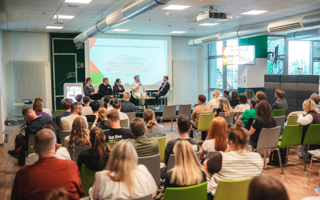 Panel of five people sitting on stage during a presentation, with a projected slide and audience in a bright, modern conference room with green chairs.