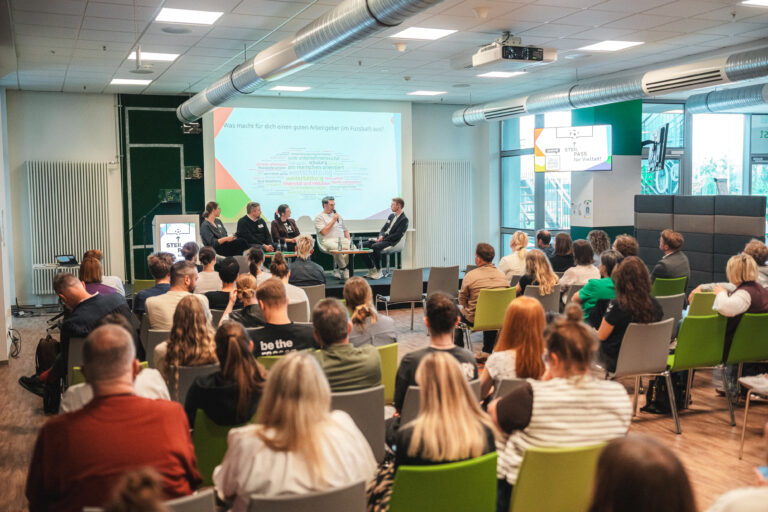 Panel of five people sitting on stage during a presentation, with a projected slide and audience in a bright, modern conference room with green chairs.