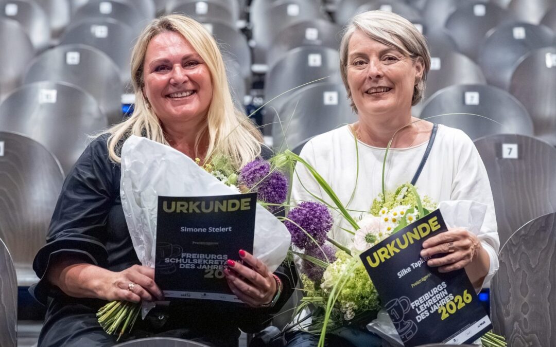 Two smiling women holding 'URKUNDE' certificates with bouquets in a stadium seating area behind them.