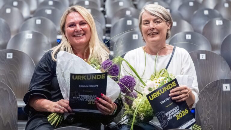 Two smiling women holding 'URKUNDE' certificates with bouquets in a stadium seating area behind them.