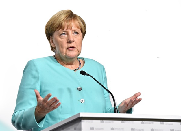 Woman in a turquoise blazer speaks at a podium with a microphone, gesturing with her hands.