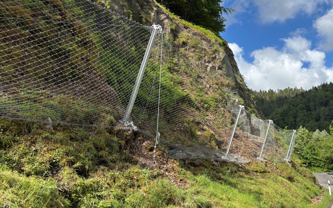 Rockfall protection fencing along a rocky hillside, with mesh netting and metal posts, beside a road.