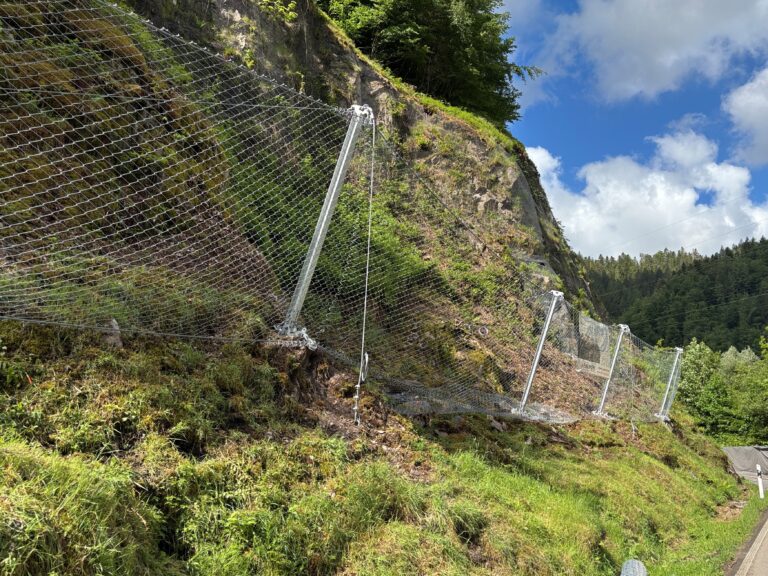 Rockfall protection fencing along a rocky hillside, with mesh netting and metal posts, beside a road.