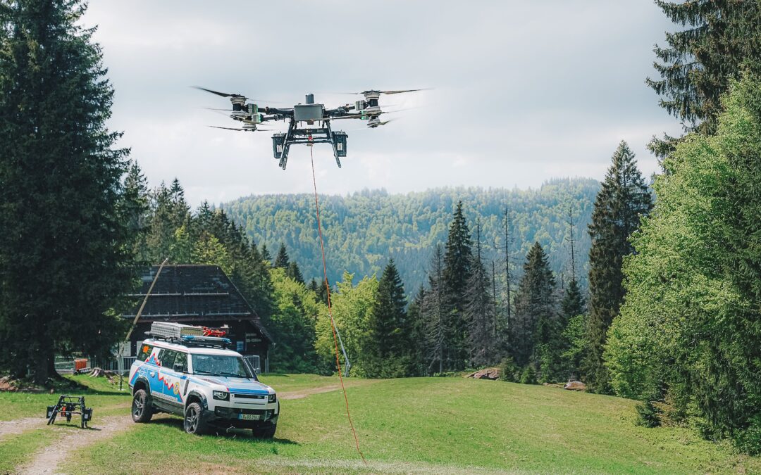 Large quadcopter drone hovering over a grassy field with an orange tether trailing downward and a support van parked nearby in a forested area.