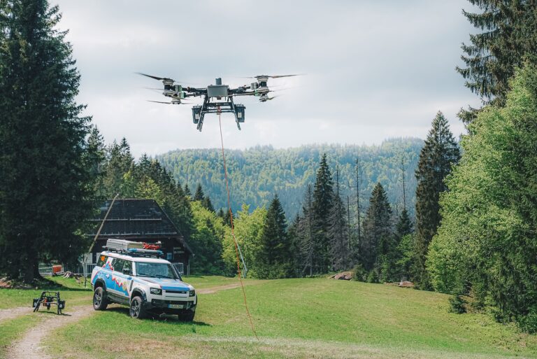 Large quadcopter drone hovering over a grassy field with an orange tether trailing downward and a support van parked nearby in a forested area.
