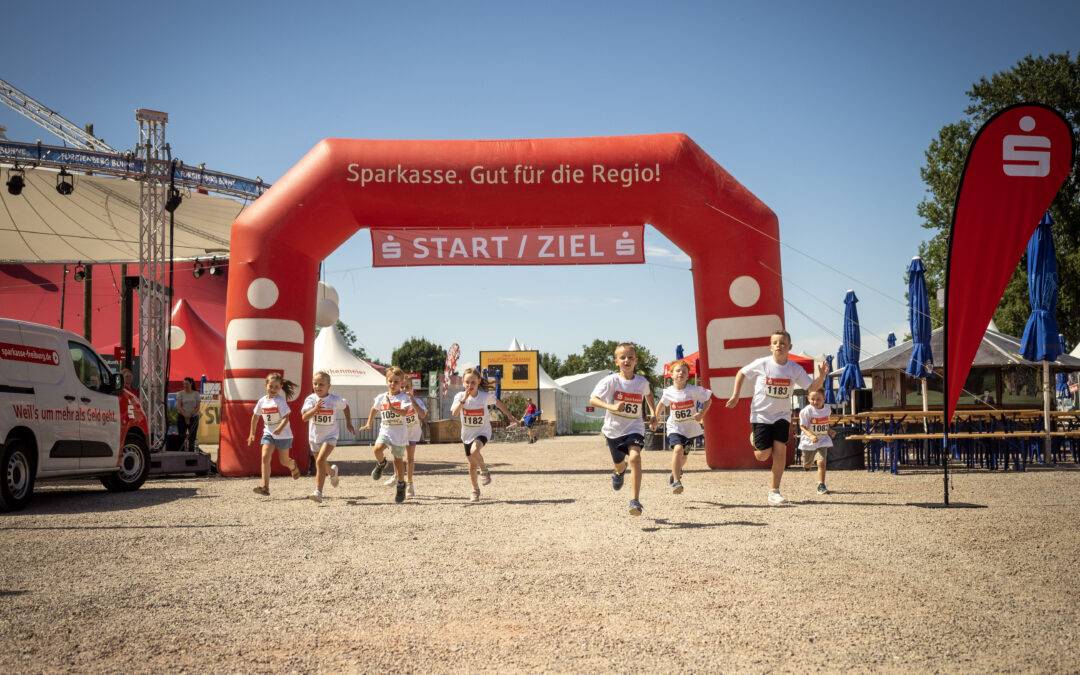 Young runners race under a red start arch labeled START/ZIEL at a daytime outdoor event.