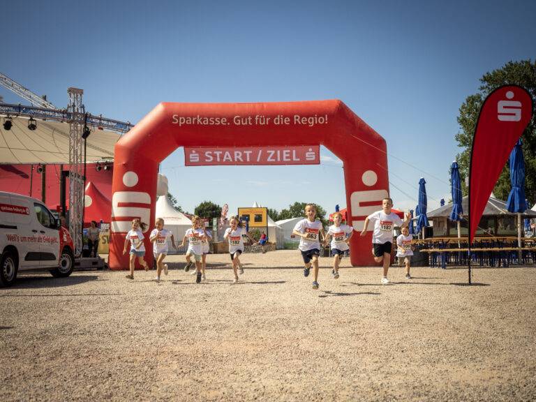 Young runners race under a red start arch labeled START/ZIEL at a daytime outdoor event.