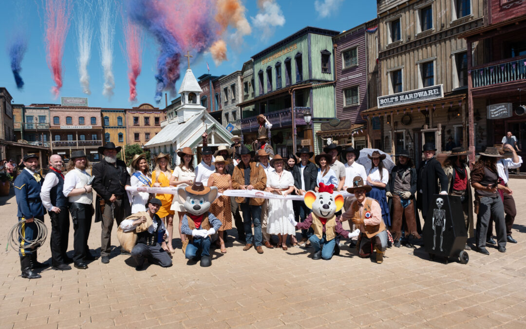 Group of people in cowboy and vintage costumes pose for a photo in a Western town set, with color smoke trails in the sky above them.