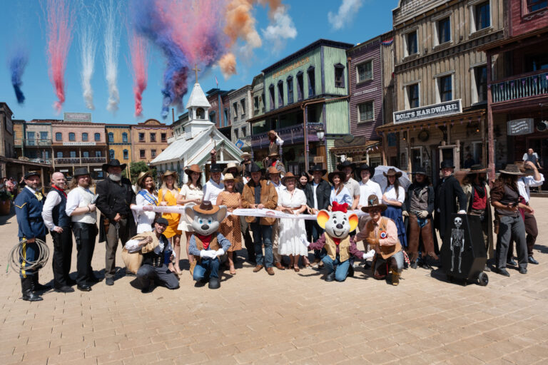 Group of people in cowboy and vintage costumes pose for a photo in a Western town set, with color smoke trails in the sky above them.