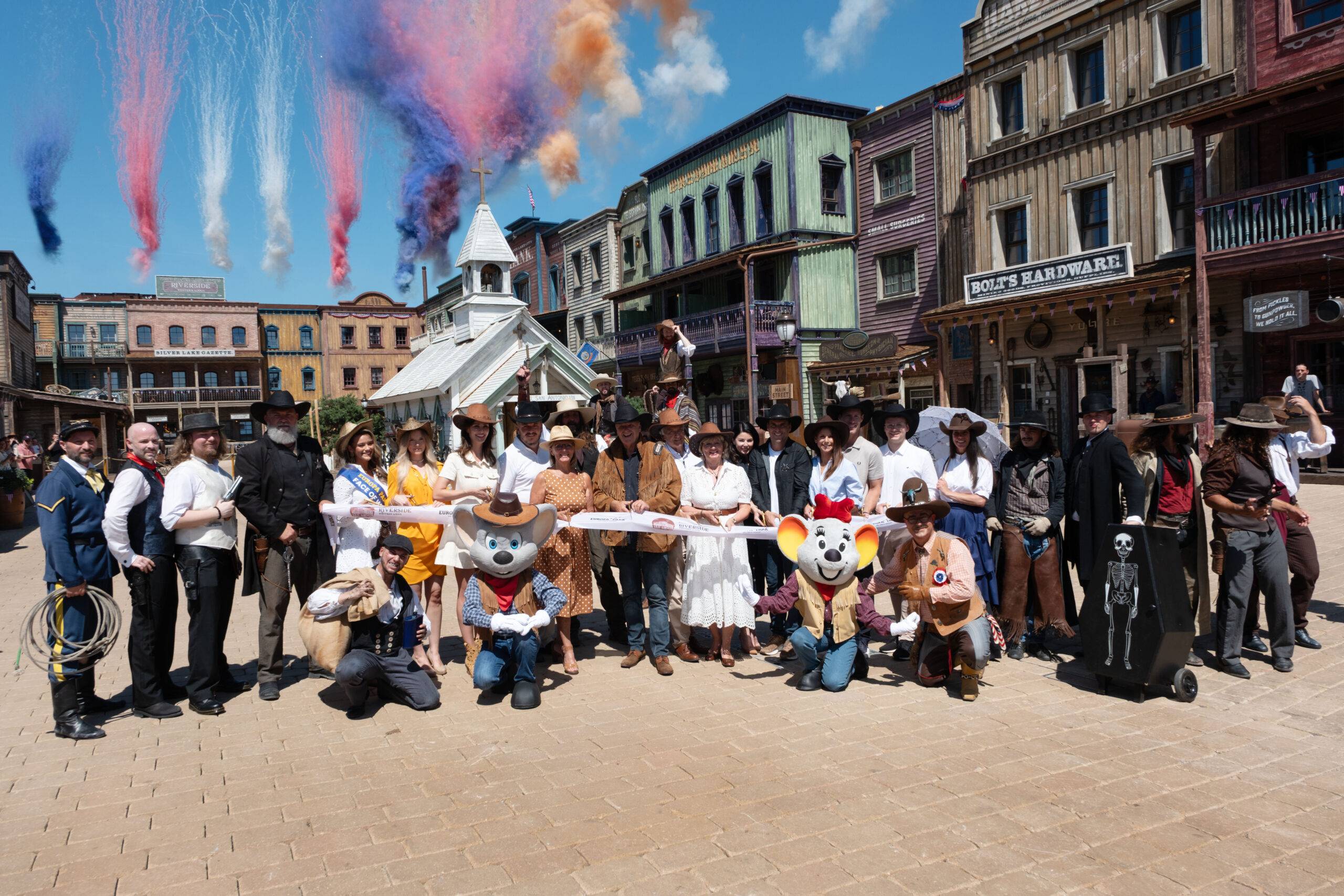 Group of people in cowboy and vintage costumes pose for a photo in a Western town set, with color smoke trails in the sky above them.