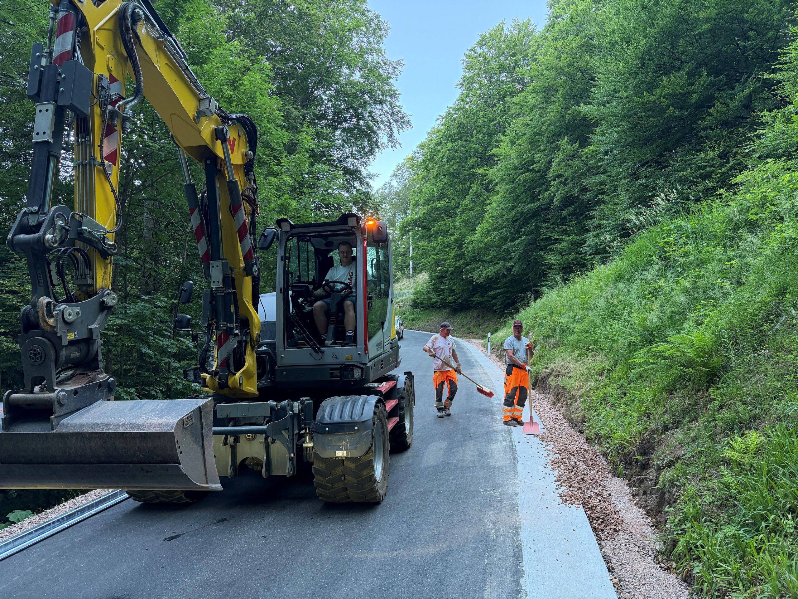 Yellow hydraulic excavator on a newly paved road beside a forest, with two workers in orange safety gear nearby.