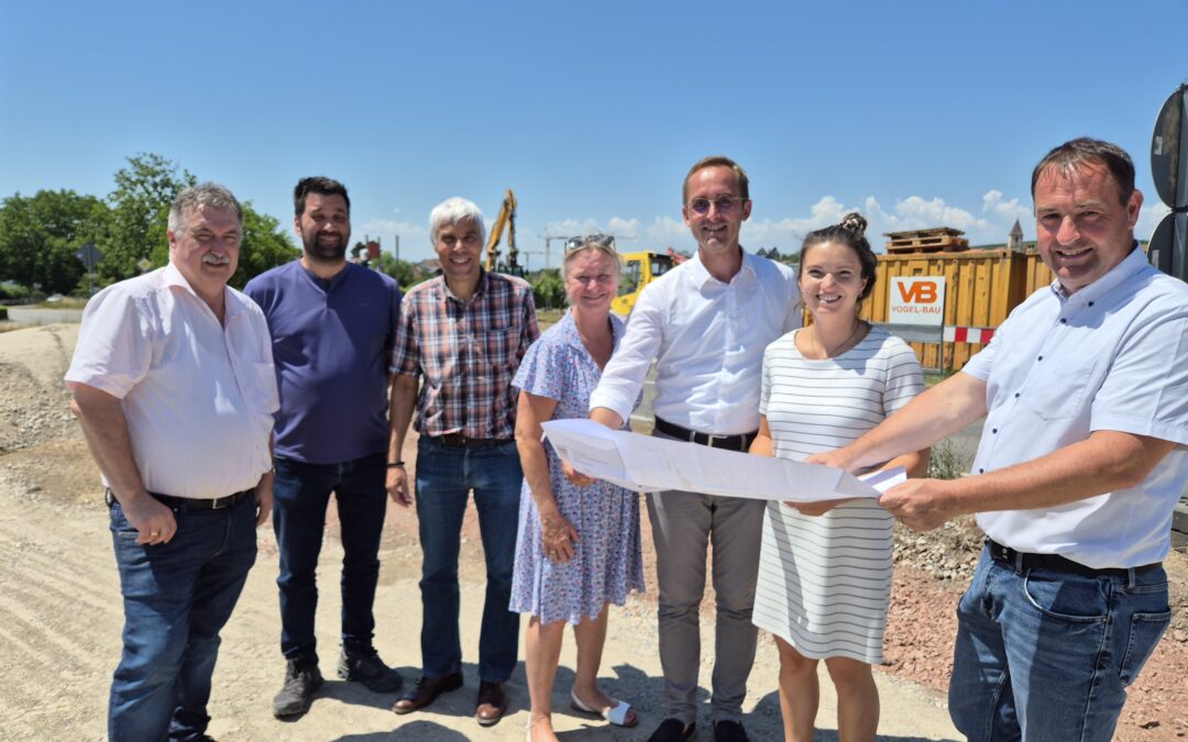 Group of six adults at a construction site, holding large blueprints and smiling at the camera, with equipment and dirt ground in the background.