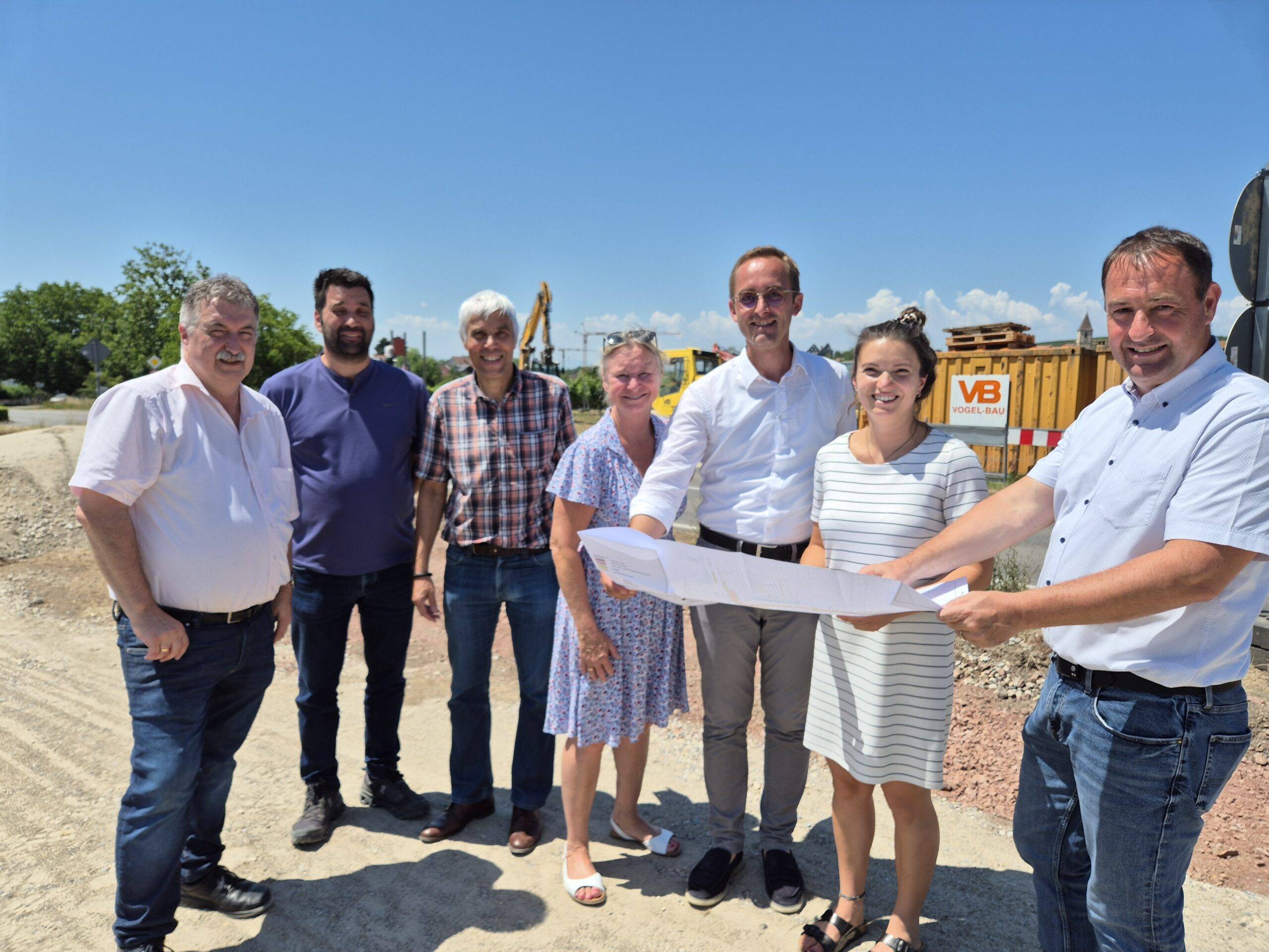 Group of six adults at a construction site, holding large blueprints and smiling at the camera, with equipment and dirt ground in the background.