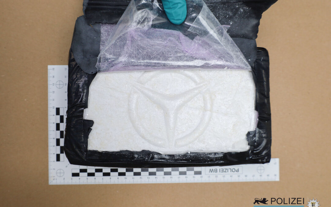 Clear plastic bag containing white powder placed on a black evidence bag, with a gloved hand at the top, measuring rulers along the sides and a police badge in the corner indicating seized evidence.