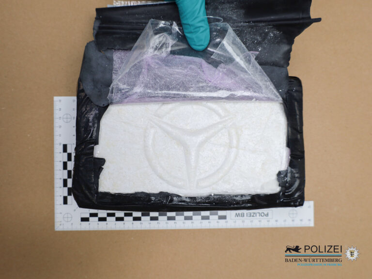 Clear plastic bag containing white powder placed on a black evidence bag, with a gloved hand at the top, measuring rulers along the sides and a police badge in the corner indicating seized evidence.