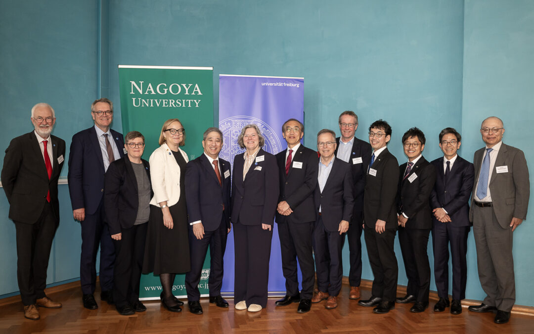 Group of professionals in formal attire posing for a photo in front of banners reading Nagoya University and Universität Freiburg.