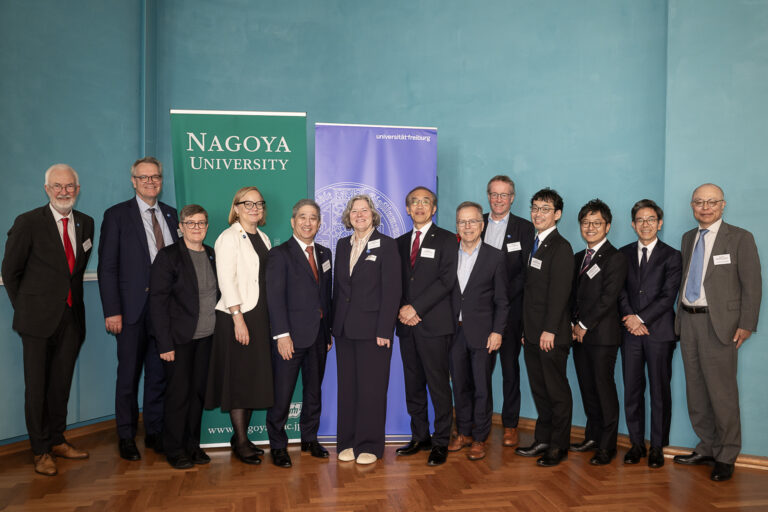 Group of professionals in formal attire posing for a photo in front of banners reading Nagoya University and Universität Freiburg.