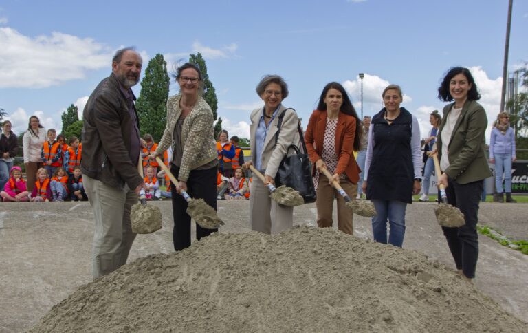 Adults and children at a groundbreaking ceremony, shovels in handle over a dirt mound, with a crowd of kids in orange vests in the background.