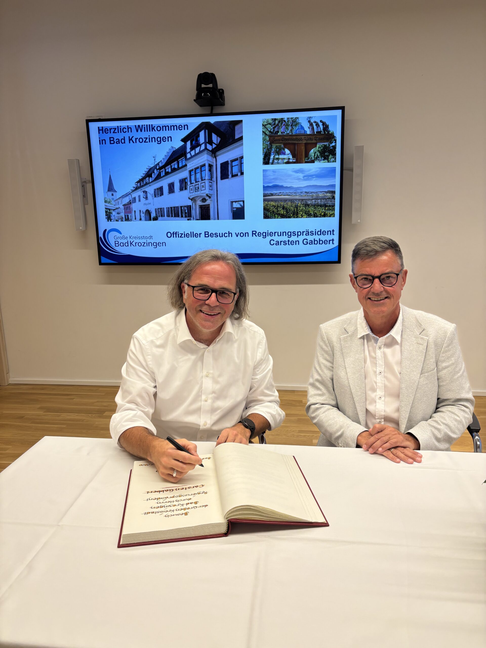 Two men in white shirts sit at a table signing a large book during an official meeting; a presentation screen behind them shows a welcome slide for Bad Krozingen.