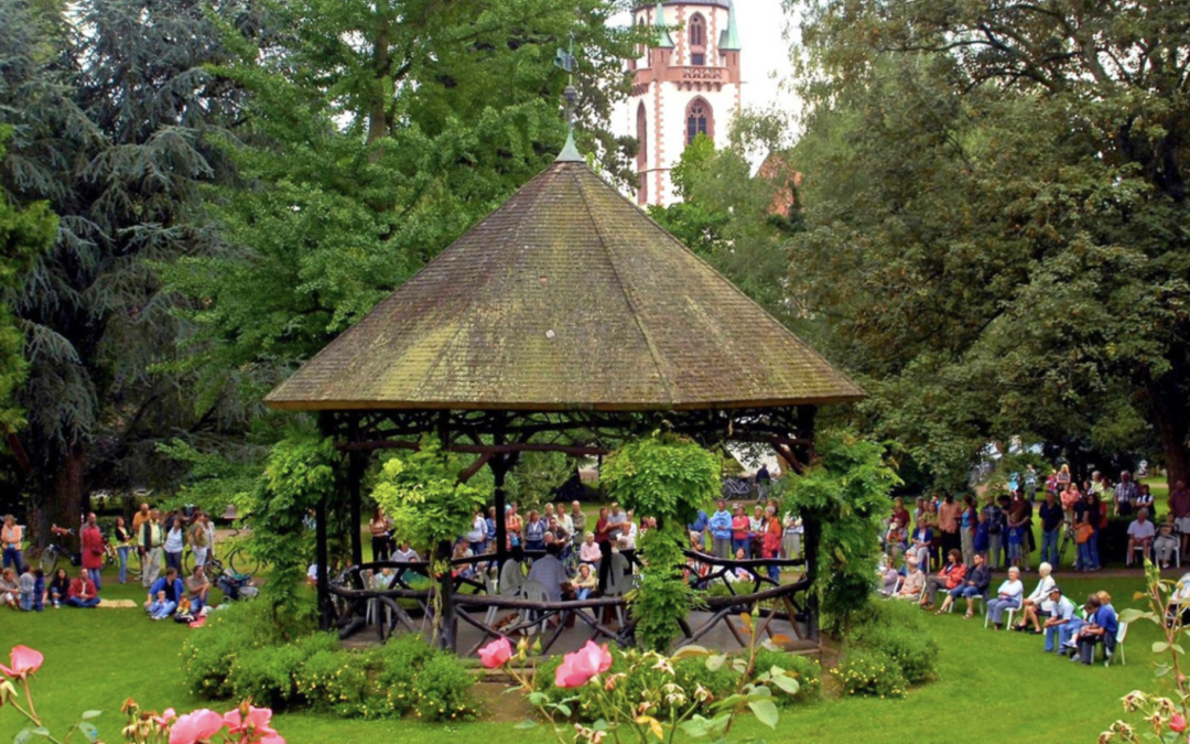 Crowd of people gathered in a park around a central thatched-roof gazebo, with trees and pink flowers in the foreground.