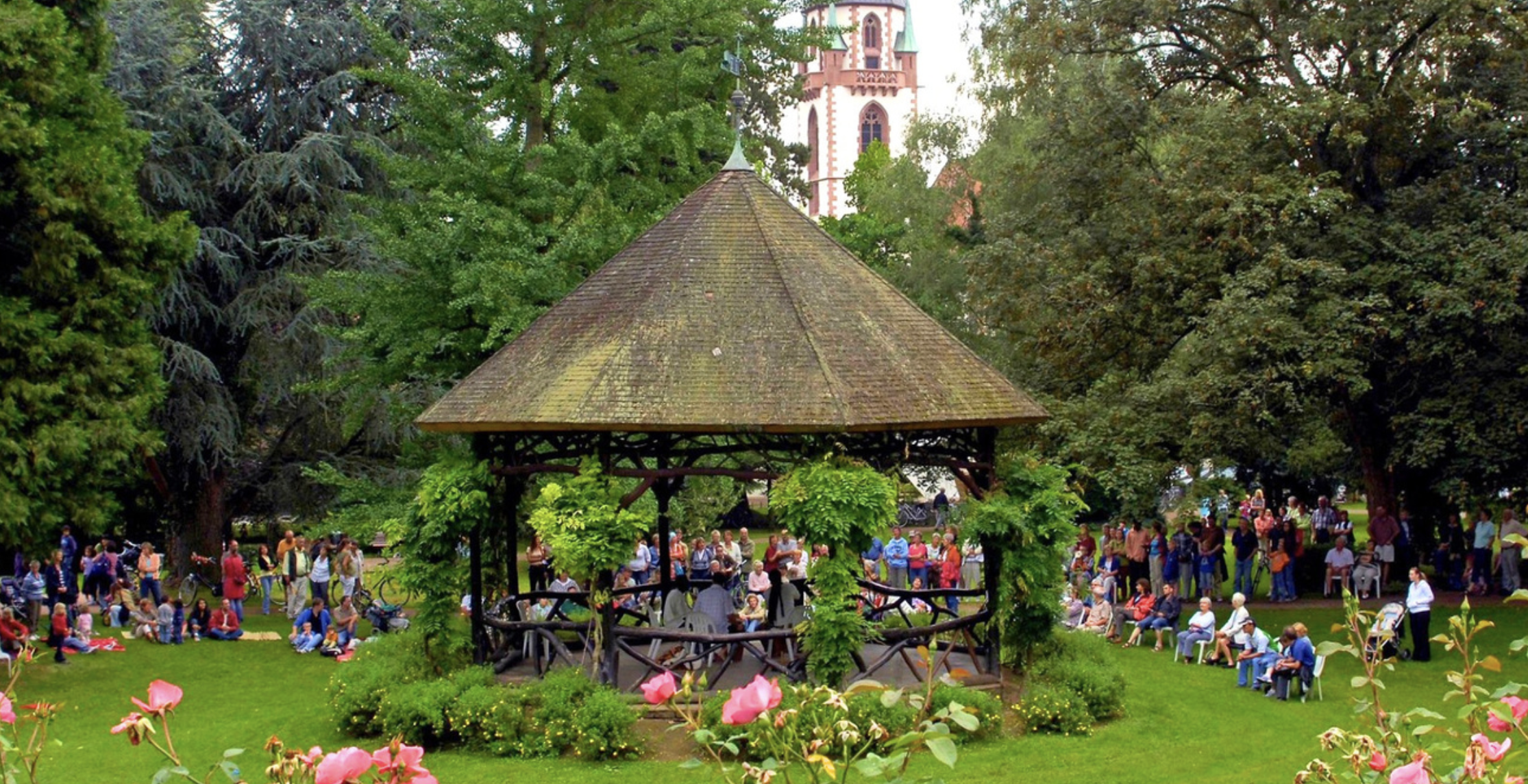Crowd of people gathered in a park around a central thatched-roof gazebo, with trees and pink flowers in the foreground.