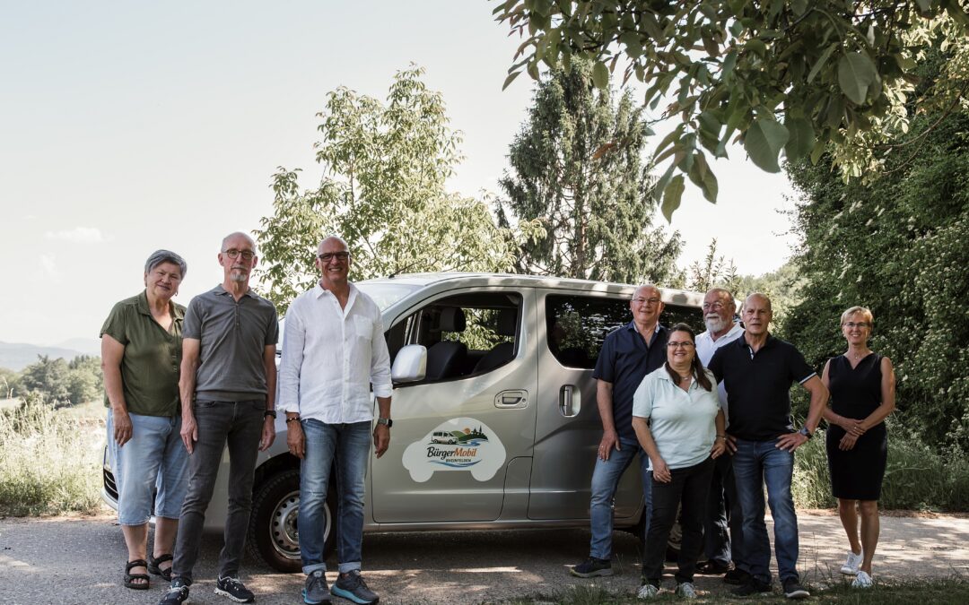 Group of adults posing beside a silver van outdoors, with trees in the background under clear sky.