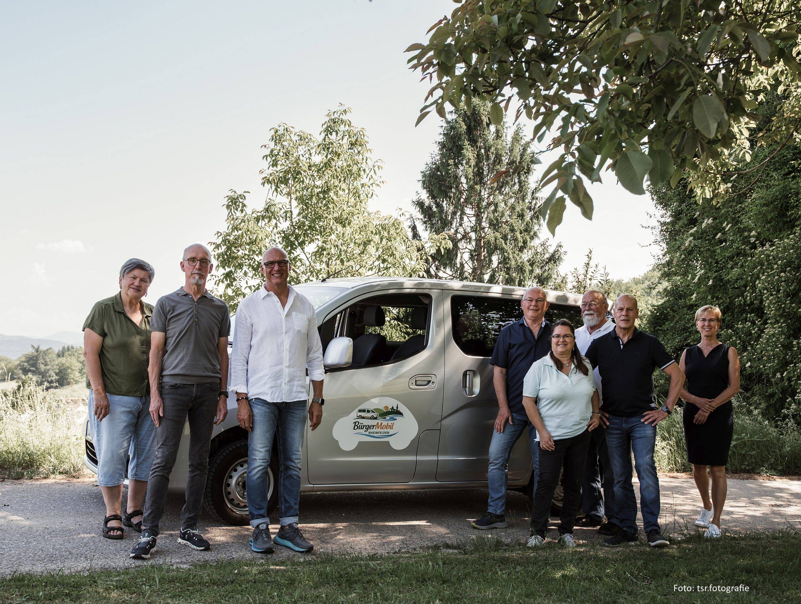 Group of adults posing beside a silver van outdoors, with trees in the background under clear sky.