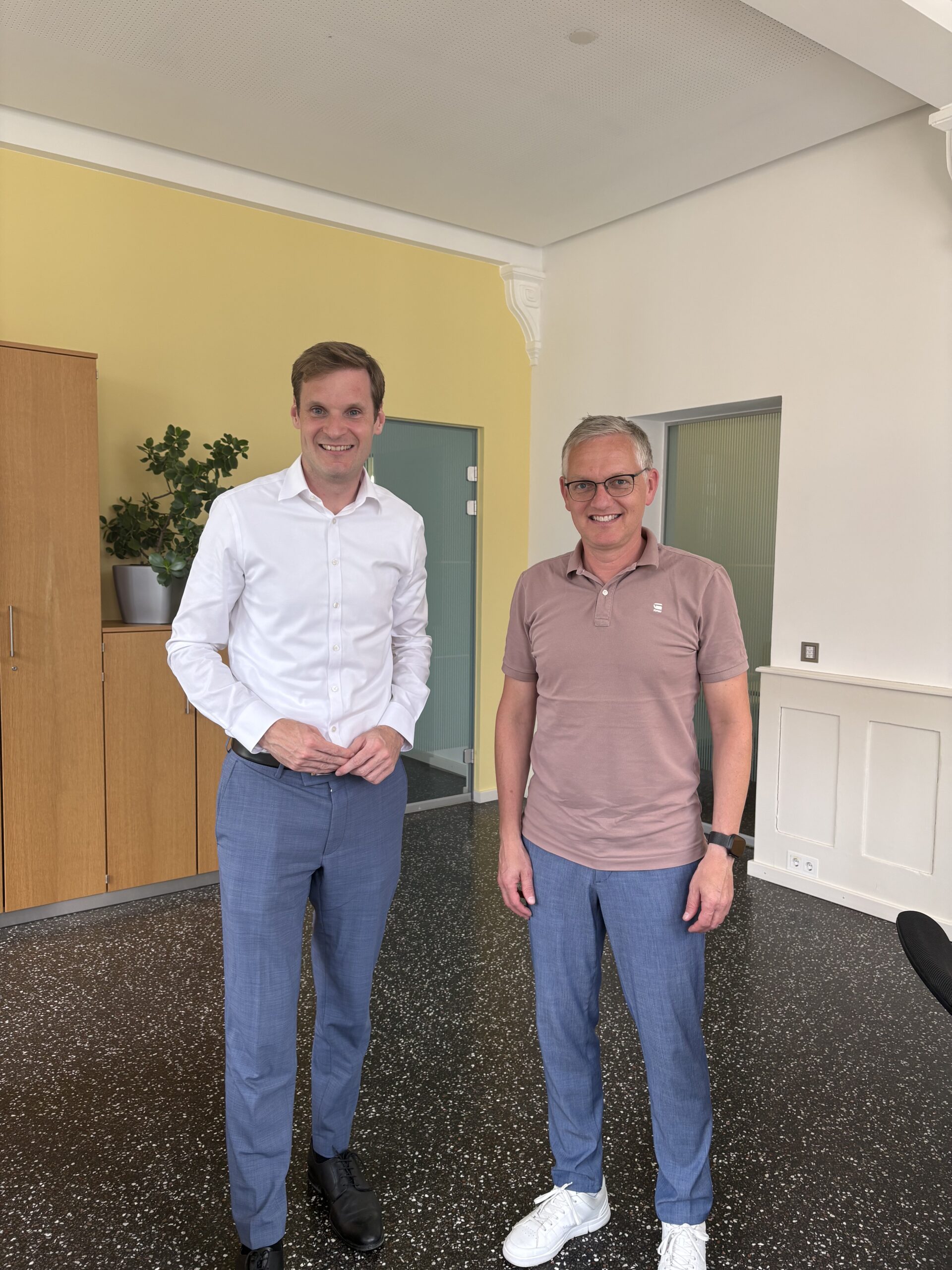 Two men stand indoors in an office space, smiling for the camera; one wears a white shirt and blue trousers, the other a pink polo and jeans with white sneakers.