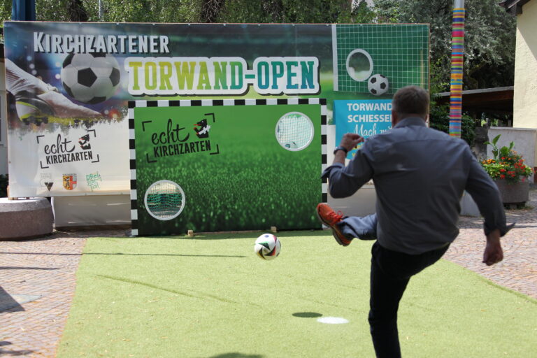 Man in a gray shirt kicks a soccer ball on an artificial turf field in front of a large advertising banner.