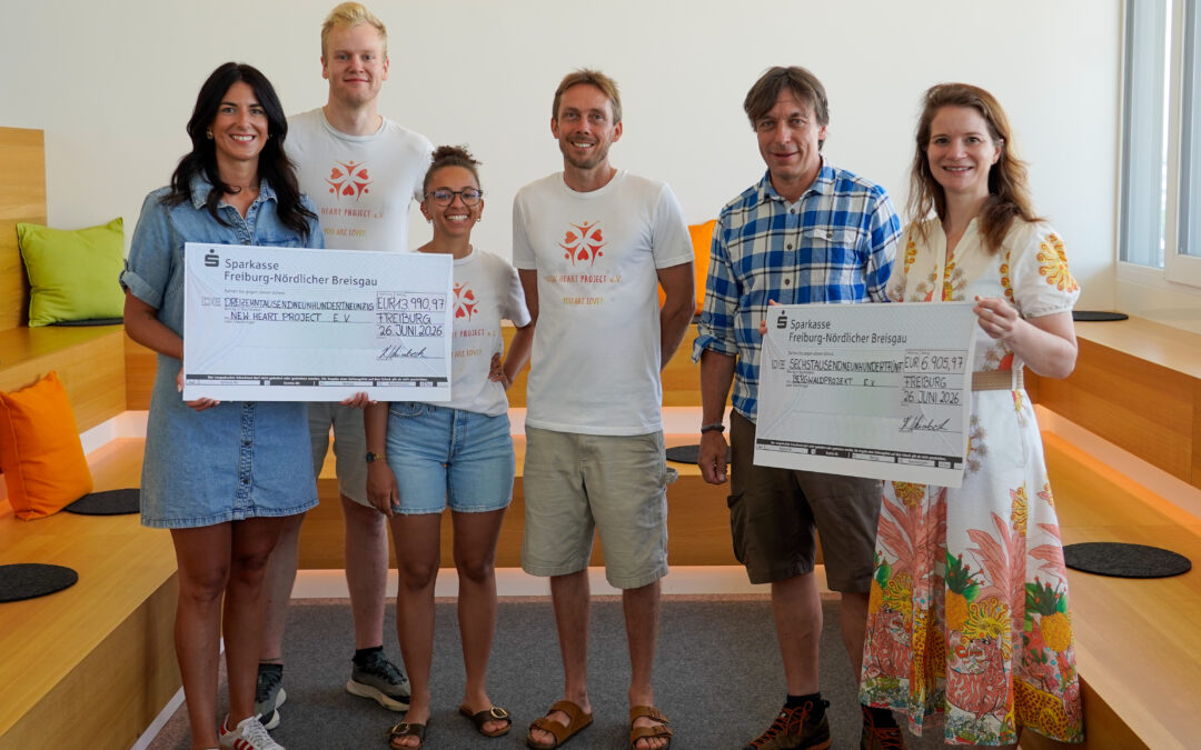 Group of six smiling people holding oversized donation checks indoors, casual summer attire.