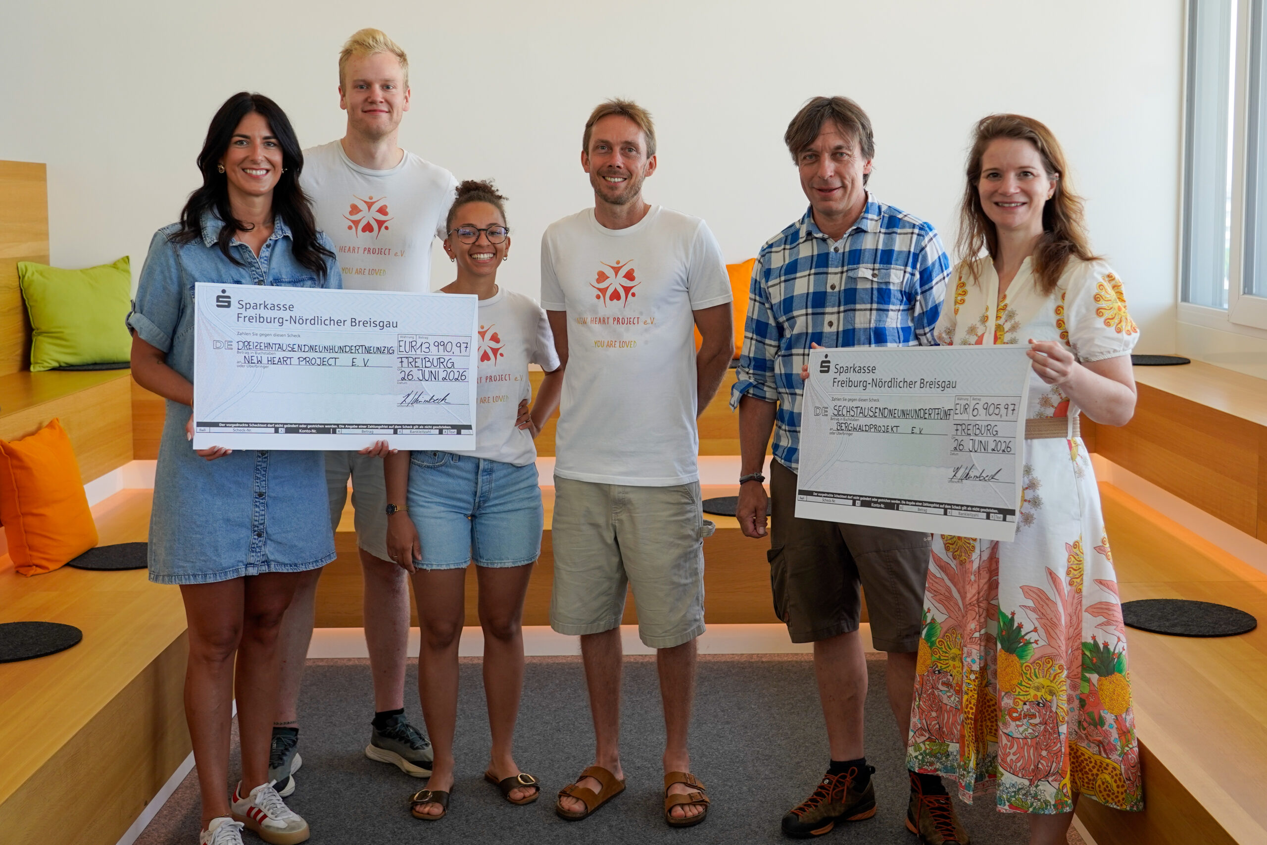 Group of six smiling people holding oversized donation checks indoors, casual summer attire.
