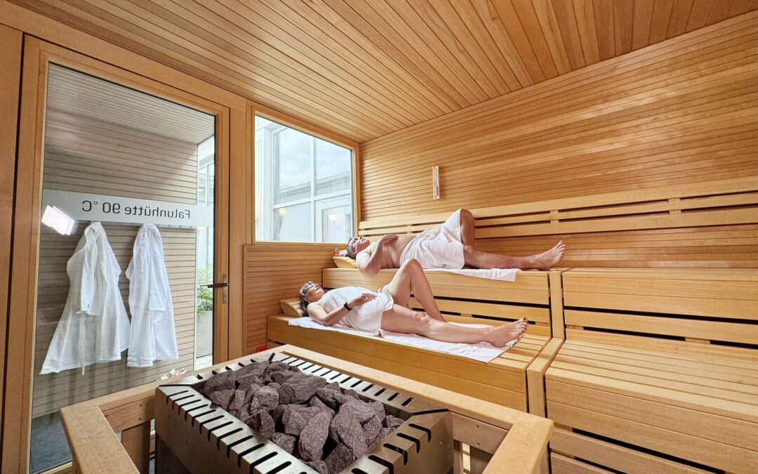 People relaxing in a wooden sauna, lying on two-tier benches with towels in a steam room salt-gray rocks on a heater in the foreground.
