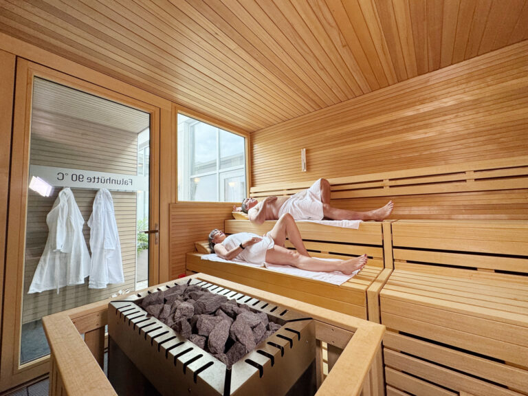 People relaxing in a wooden sauna, lying on two-tier benches with towels in a steam room salt-gray rocks on a heater in the foreground.
